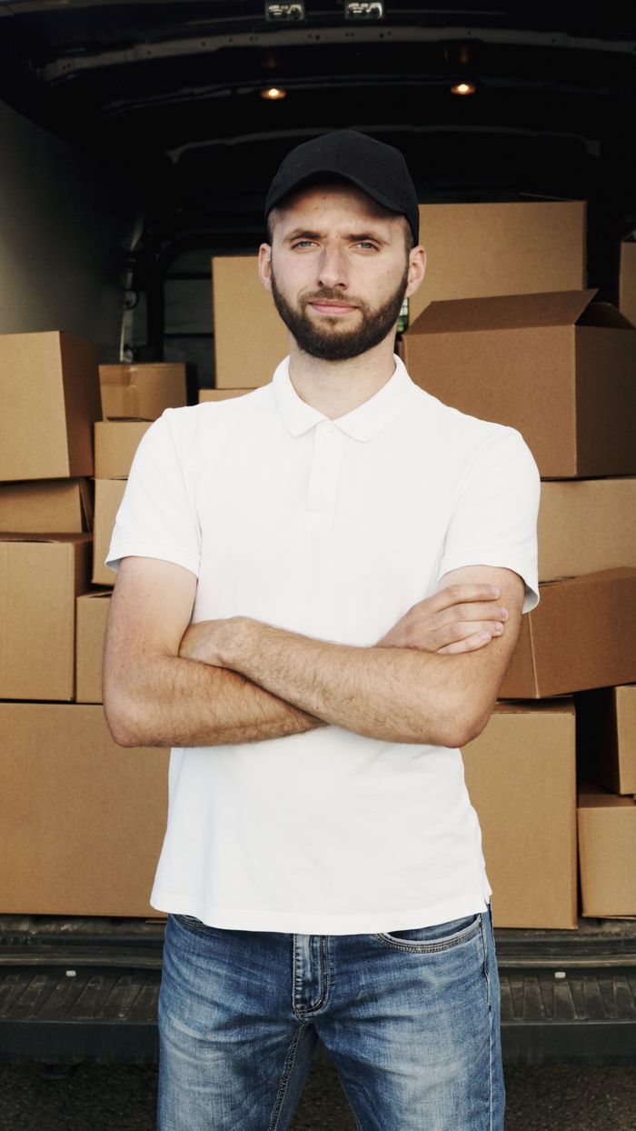 Services A deliveryman standing confidently in front of a van loaded with cardboard boxes, ready for dispatch.